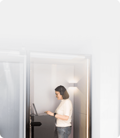 Frau mit Headset arbeitet am Laptop in einer gläsernen Telefonkabine