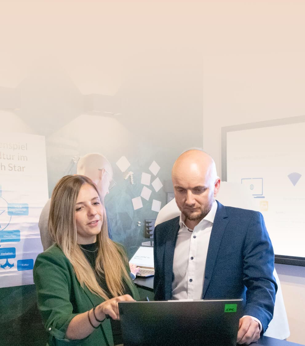 Beraterin geht mit einem Kollegen am Laptop die nächsten Schritte durch, dahinter Workshop-Plakate