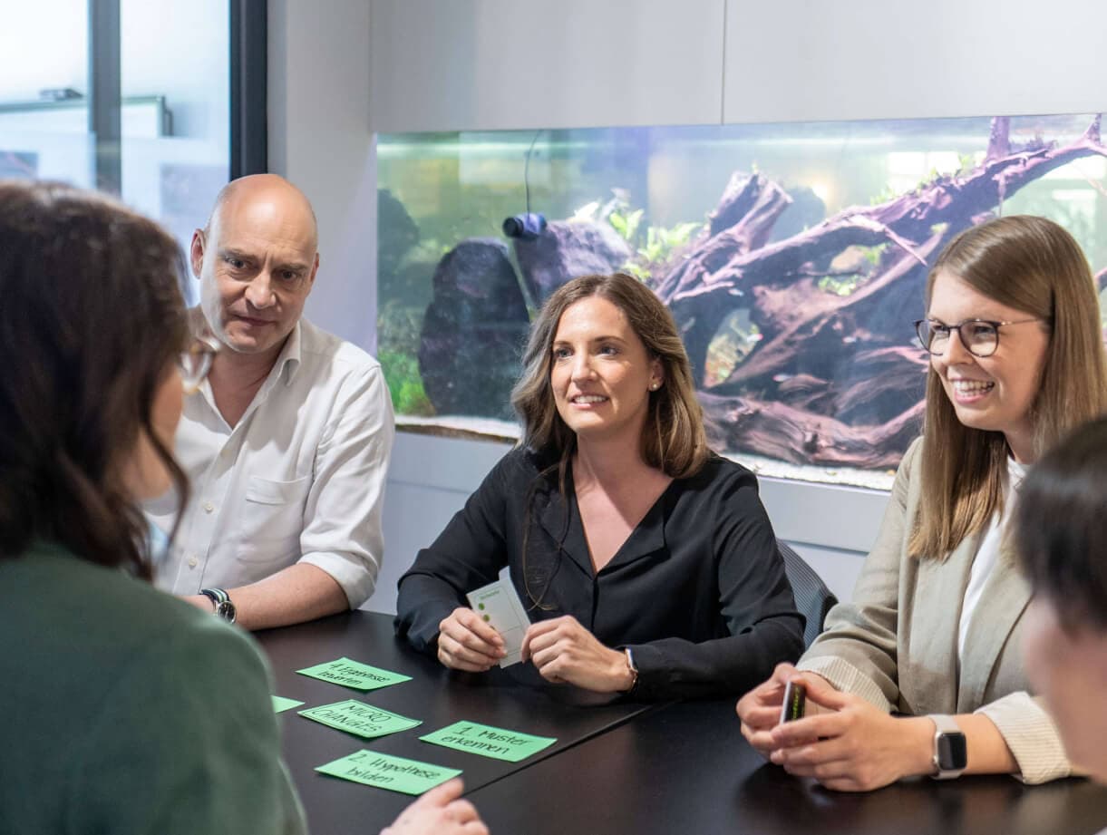 Workshop-Runde am Tisch, auf dem grüne Karten mit Schritten wie „Muster erkennen“ und „Hypothese bilden“ liegen