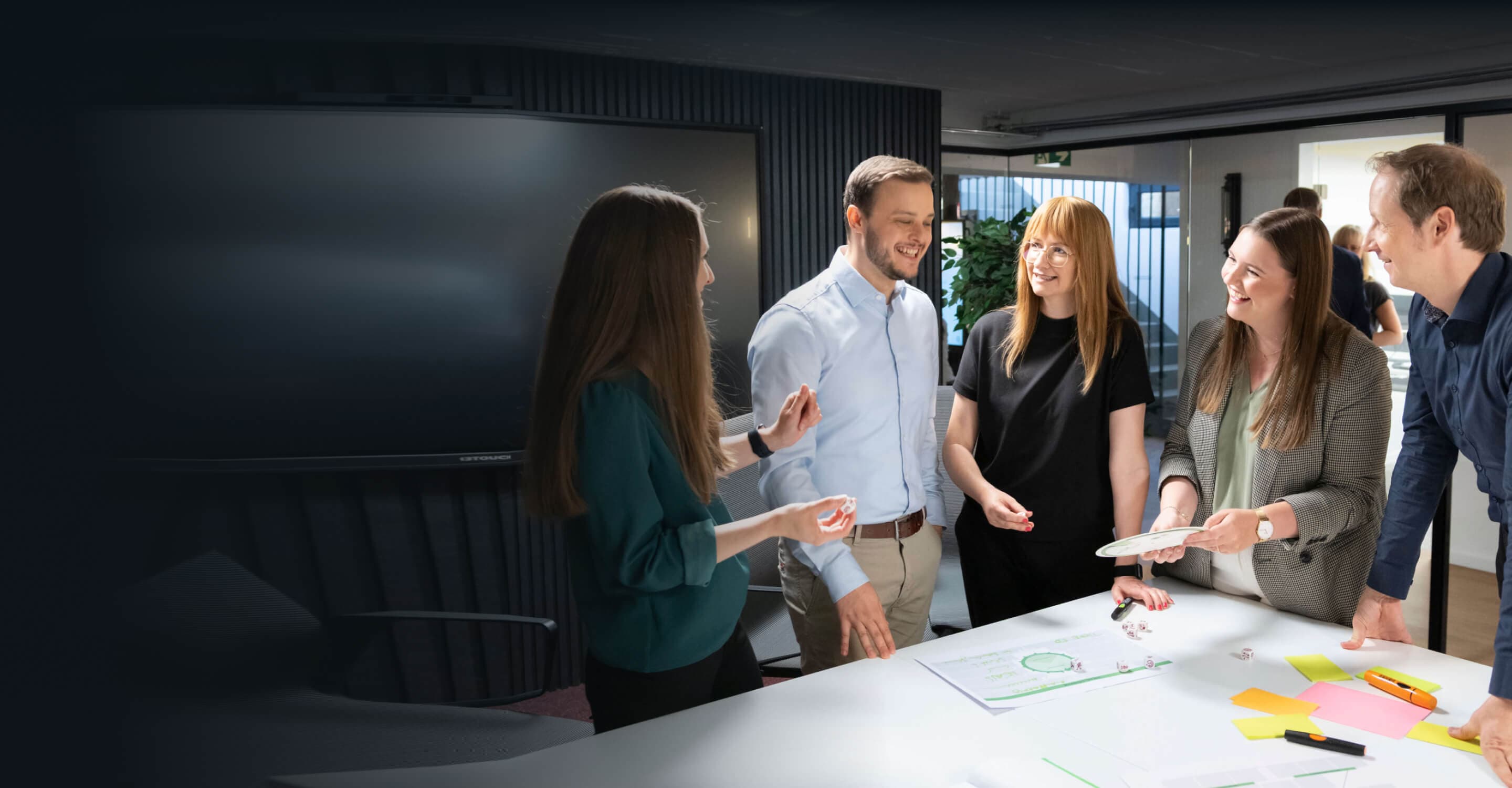 Team steht lachend um einen Tisch mit Haftnotizen und Würfeln und arbeitet im Workshop an Ideen