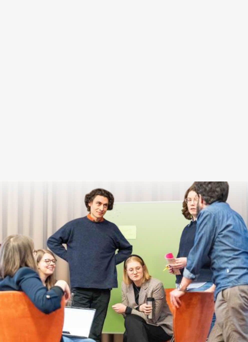 Workshop-Gruppe im Austausch, sitzend und stehend vor einer grünen Pinnwand