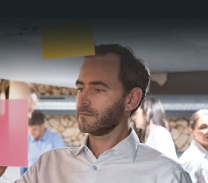 Berater klebt Haftnotizen an eine Glaswand, im Hintergrund arbeitet das Team an einem Veränderungsprojekt
