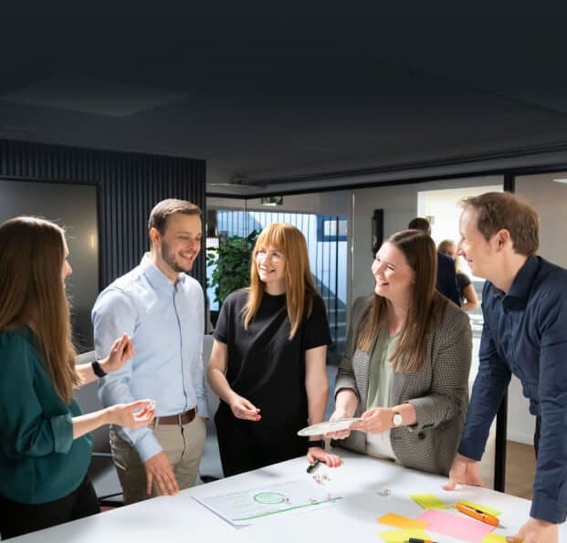 Team steht lachend um einen Tisch mit Haftnotizen und Würfeln und arbeitet im Workshop an Ideen